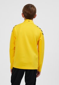 Gul sportjacka med hög krage, med svarta detaljer på axlarna. Slät textur, figursydd design och långa ärmar.