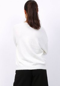 Femme aux longs cheveux noirs attachés, portant un pull blanc et un pantalon noir, se tenant de dos contre un fond blanc uni.