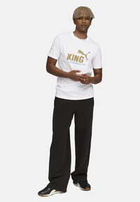 Hvid T-shirt med korte ærmer med guld- og blå tekst "KING MARSEILLE," parret med sorte brede bukser og sorte sneakers.
