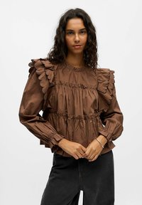 Blouse marron à volants avec des bords festonnés, design en couches, poignets élastiques et encolure ronde. Portée avec un jean noir taille haute.