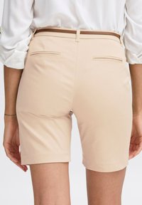 Beige nadrágos rövidnadrág sima textúrával, közepes derékkal, övlyukakkal és két hátsó zsebbel. Letisztult fazon, egyenes lábbal.