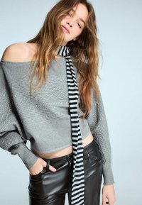 Pulover gri, texturat, cu un design off-shoulder și modele tricotate în cablu, împreună cu pantaloni din piele negri și o eșarfă cu dungi alb-negru.