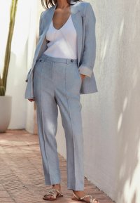 Blazer ajusté bleu clair et pantalon assorti en tissu léger, associé à un débardeur blanc à côtes et des sandales plates.