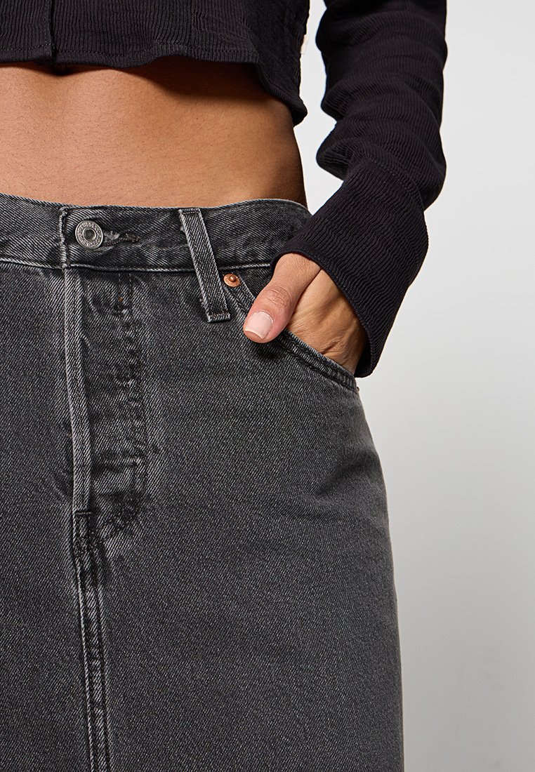 Vaqueros de mezclilla negra de cintura alta, con cierre de botón, bolsillo frontal y detalles de costura sutiles, combinados con una blusa negra de manga larga texturizada.