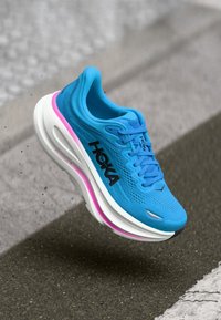 Chaussure de sport bleue avec une tige en maille, semelle intermédiaire blanche et accents roses. Présente une forme arrondie et un logo proéminent sur le côté.