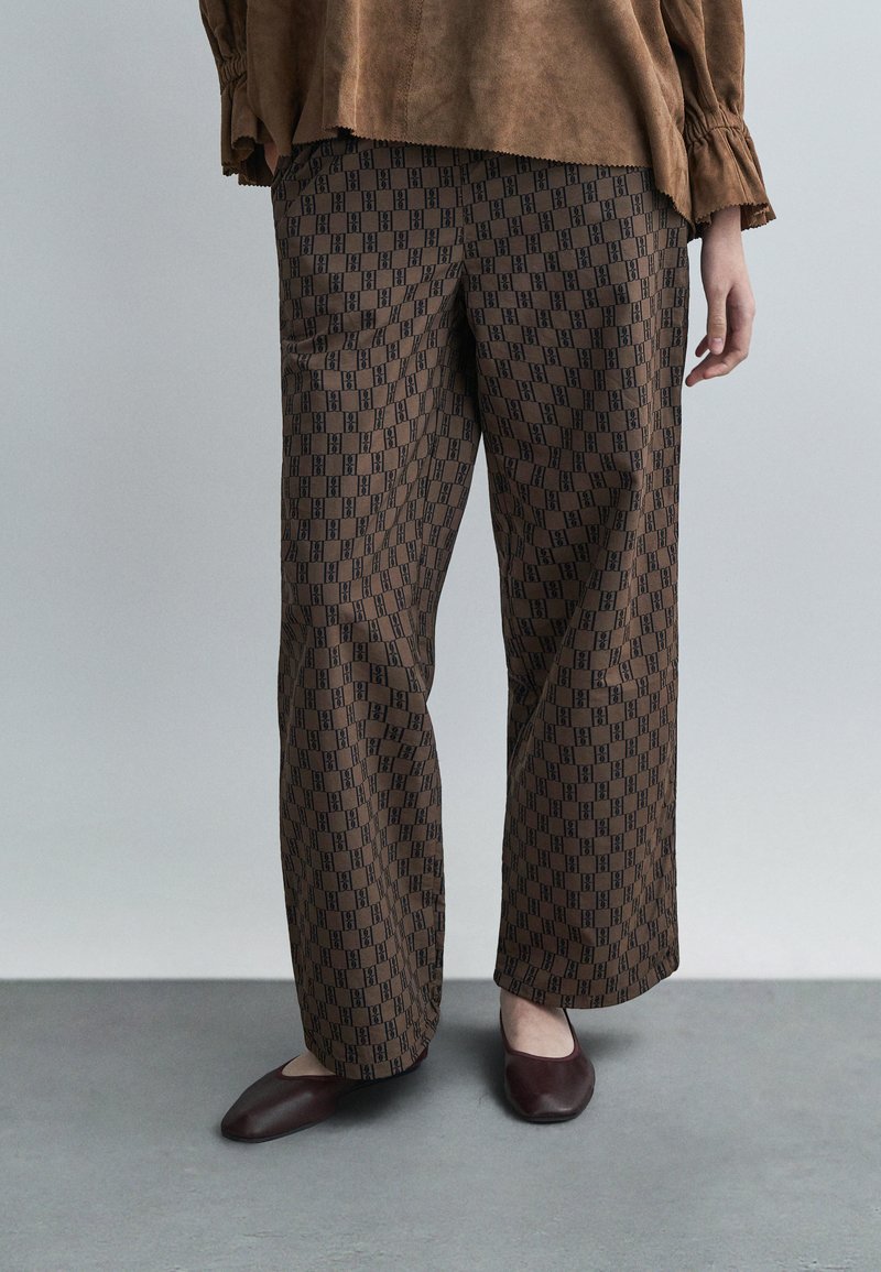 Person iført brune mønstrede bukser med vide ben og matchende brune flate sko, stående på et grått gulv mot en ensfarget bakgrunn.