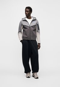 Pelēka un melna rāvējslēdzēja džemperis ar kapuci, kontrastējošām piedurknēm un nelielu zilu logotipu. Valdziņā ar vaļīgām melnām biksēm un pelēkām kedām.