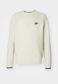 Krämfärgad Nike-tröja med svart logotyp, ribbad halsringning och manschetter samt raglansärmar. Slät textur, avslappnad design.