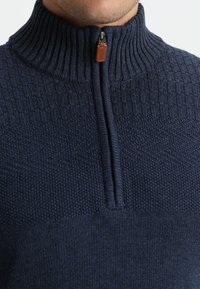 Tummansininen vetoketjupaita, jossa on korkea kaulus. Yläosassa on teksturoitu neulekuvio ja alaosassa sileä pinta. Vetoketjullinen etuosa, jossa on nahkakoristelu.