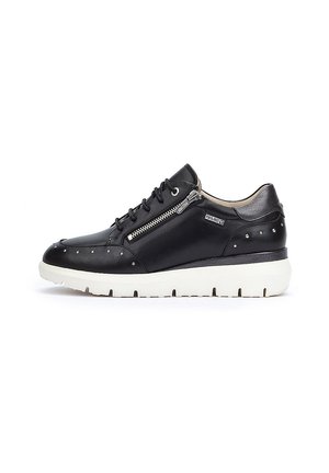 Zapatilla de cuero negra con suela de goma blanca, que presenta un cierre lateral, acentos con tachuelas y un diseño de punta redondeada. Textura suave en toda la superficie.