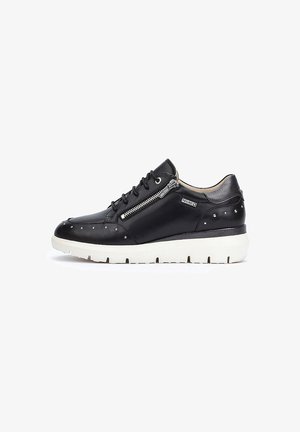 Zapatilla de cuero negra con suela de goma blanca, que presenta un cierre lateral, acentos con tachuelas y un diseño de punta redondeada. Textura suave en toda la superficie.