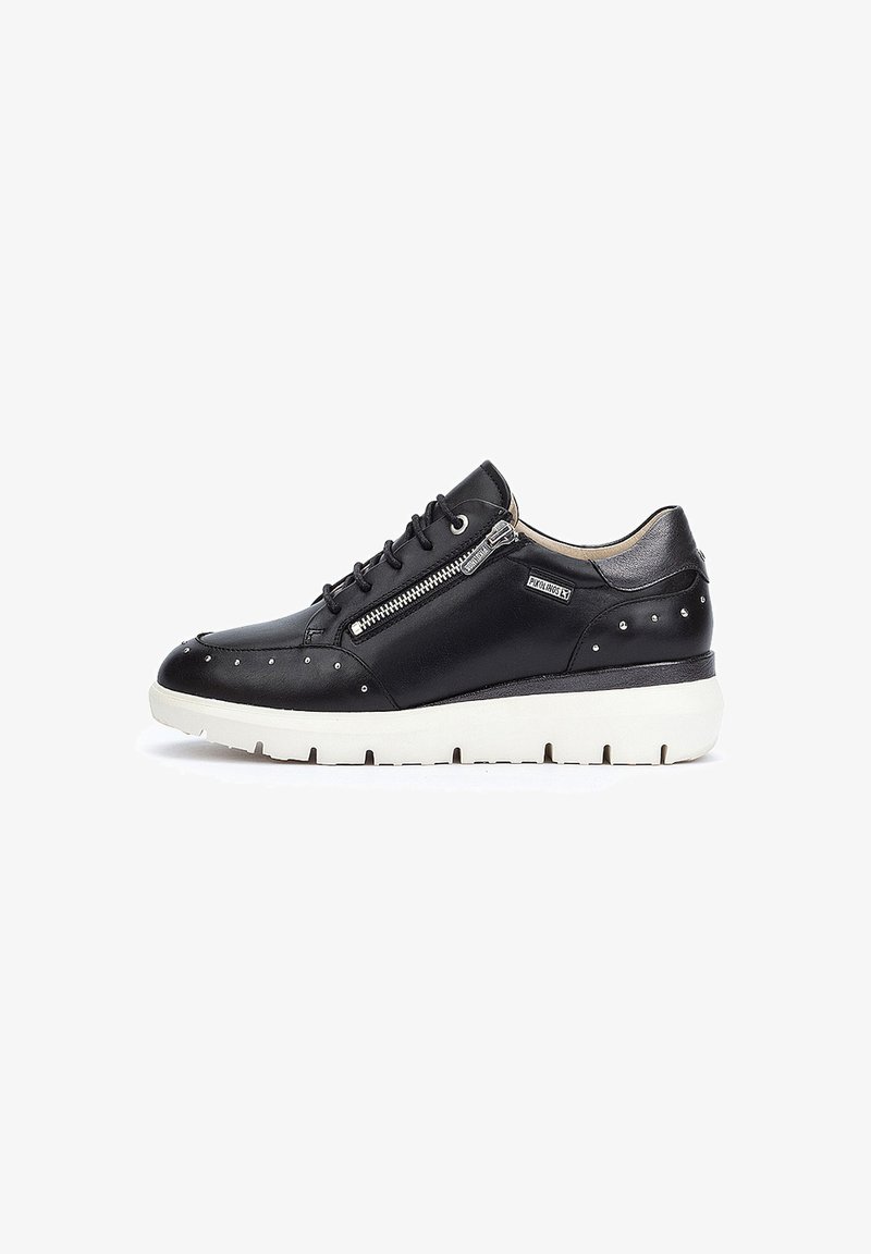 Zapatilla de cuero negra con suela de goma blanca, que presenta un cierre lateral, acentos con tachuelas y un diseño de punta redondeada. Textura suave en toda la superficie.