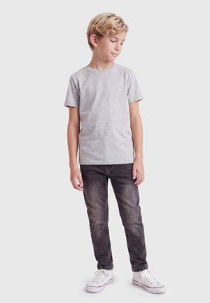Garçon aux cheveux blonds debout, regardant vers le bas, portant un t-shirt gris clair, un jean noir délavé et des baskets blanches sur un fond blanc uni.
