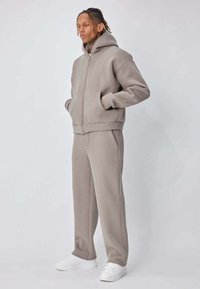 Personne portant un sweat à capuche taupe zippé et un pantalon large assorti, debout les mains dans les poches, chaussures blanches aux pieds.