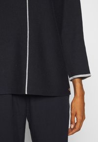 Granatowy, dziergany top z długimi rękawami, z kontrastowym białym paskiem wzdłuż środka i jasnoszarym mankietem. Złote detale z okuciami na dole.