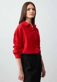 Rotes Leinenhemd mit Kragen, Knopfverschluss vorne und langen, hochgekrempelten Ärmeln. Kombiniert mit einem schwarzen Rock für den Kontrast.