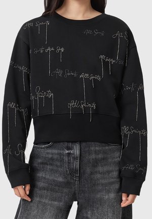 Sweat-shirt court noir avec texte argenté brodé "All Saints" répété et jean gris foncé délavé porté par une personne aux longs cheveux foncés.