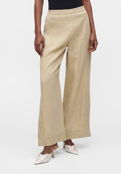 Pantalon beige à jambes larges avec des broderies discrètes près des ourlets, porté avec des mules blanches à talons et un haut noir.