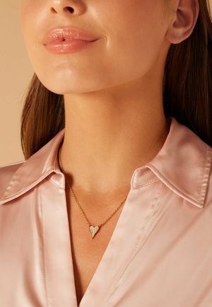 Gouden ketting met een hartvormige hanger met een gestructureerd oppervlak en sprankelende details, gedragen over een lichtroze satijnen shirt.