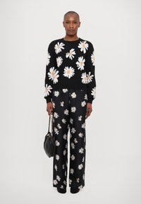 Schwarzer Pullover mit weißer und gelber Gänseblümchen-Stickerei, kombiniert mit weit geschnittenen schwarzen Hosen mit einem ähnlichen Blumenmuster. Schwarze Handtasche.