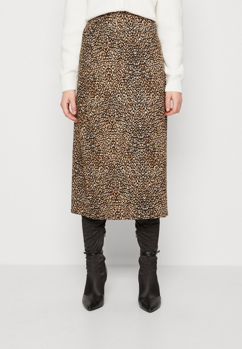 Femme portant une jupe midi à imprimé léopard, un pull blanc et des bottes noires à talons pointus, debout devant un fond blanc.
