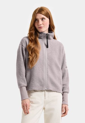 Young woman with long red hair wearing a light purple fleece jacket with a high collar and off-white pants, standing against a plain background.