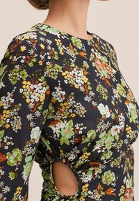 Květinové šaty z tmavé tkaniny, zdobené živými zelenými, oranžovými a bílými vzory. Obsahují kulatý výstřih a postranní výřezy pro detail.