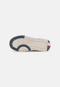 Suola della scarpa in beige, con un design di grip a motivi. Accenti in blu navy e rosso vicino al tallone. Logo del marchio visibile sulla suola.