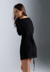 Svart kjole med en fitted bodice, lange ermer og en flytende skjørt. Har en knyttet detalj bak og gjennomsiktige aksenter langs ermene.