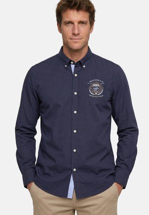 Hombre con camisa azul marino abotonada con emblema bordado en el pecho izquierdo, combinado con pantalones beige, de pie con las manos en los bolsillos.