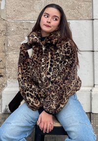 Casaco de pele sintética com padrão de leopardo, gola alta e fecho de correr, combinado com jeans azul claro. Textura suave e ajuste oversized.