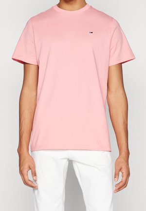 Mand iført en ensfarvet lyserød T-shirt med korte ærmer og et lille logo på brystet samt hvide bukser, stående mod en grå baggrund.