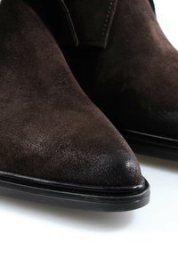 Gros plan de bottes en daim marron à bout pointu avec semelles en caoutchouc noir sur une surface blanche.