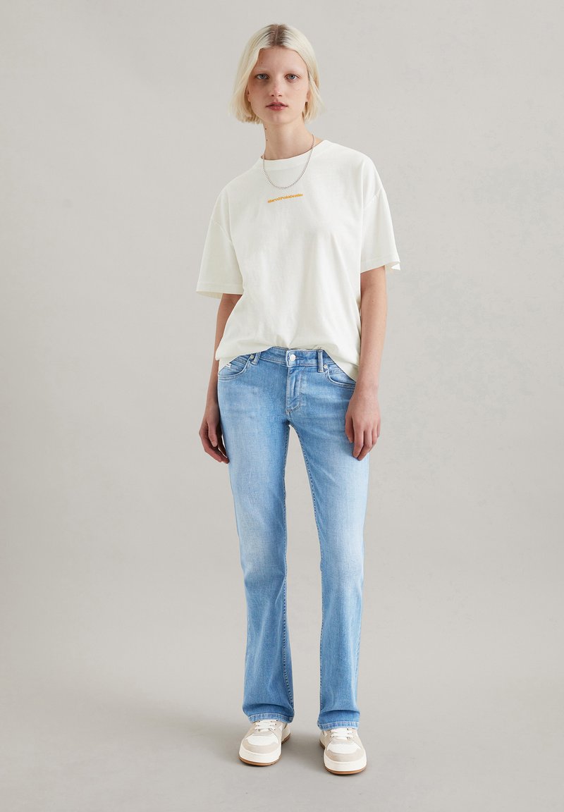 Biała, oversize'owa koszulka z pomarańczowym napisem, zestawiona z jasnoniebieskimi, rozszerzającymi się dżinsami i kremowymi trampkami. Minimalistyczny design i swobodny styl.