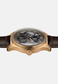 Montre squelette couleur or avec un cadran gris rond, un bracelet en cuir marron, de grands chiffres romains comme repères, et une couronne texturée sur le côté.