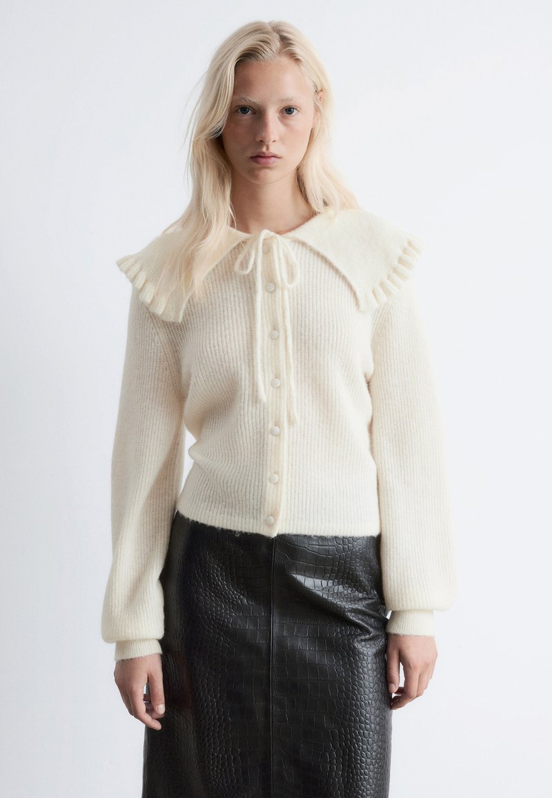 Helmfarbener Cardigan mit Rüschenkragen und gerippter Struktur, vorne mit Knöpfen, kombiniert mit einem schwarzen Kunstlederrock mit Krokodilmuster.