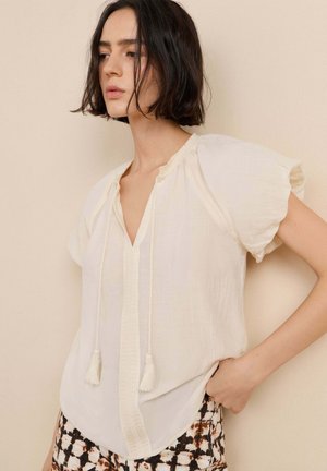 Junge Frau mit kurzen dunklen Haaren, die eine lockere weiße Bluse mit Quastenbändern und gemusterte schwarze, braune und weiße Hose trägt, steht vor einer beigen Wand.