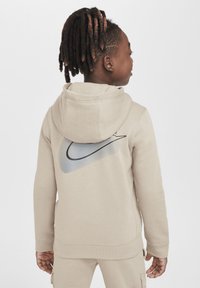 Hanorac bej cu glugă, având un logo mare negru Nike cu un gradient în spate. Textură netedă, design casual, manșete rulade.