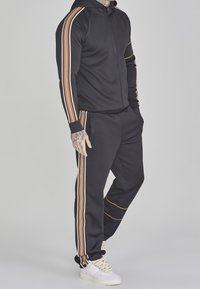 Survêtement noir avec capuche zippée, présentant des rayures verticales marron et orange sur les manches et les jambes ; tissu lisse et ceinture élastique.