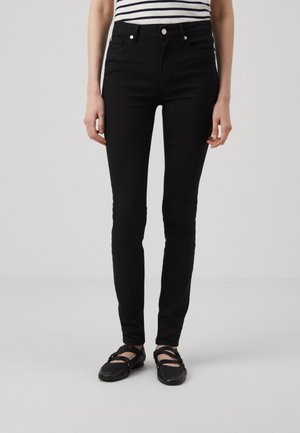 Sort smalle jeans båret med sorte stroppesko og et hvidt og sort stribet top, vist fra midt på overkroppen til fødderne på en ensfarvet baggrund.
