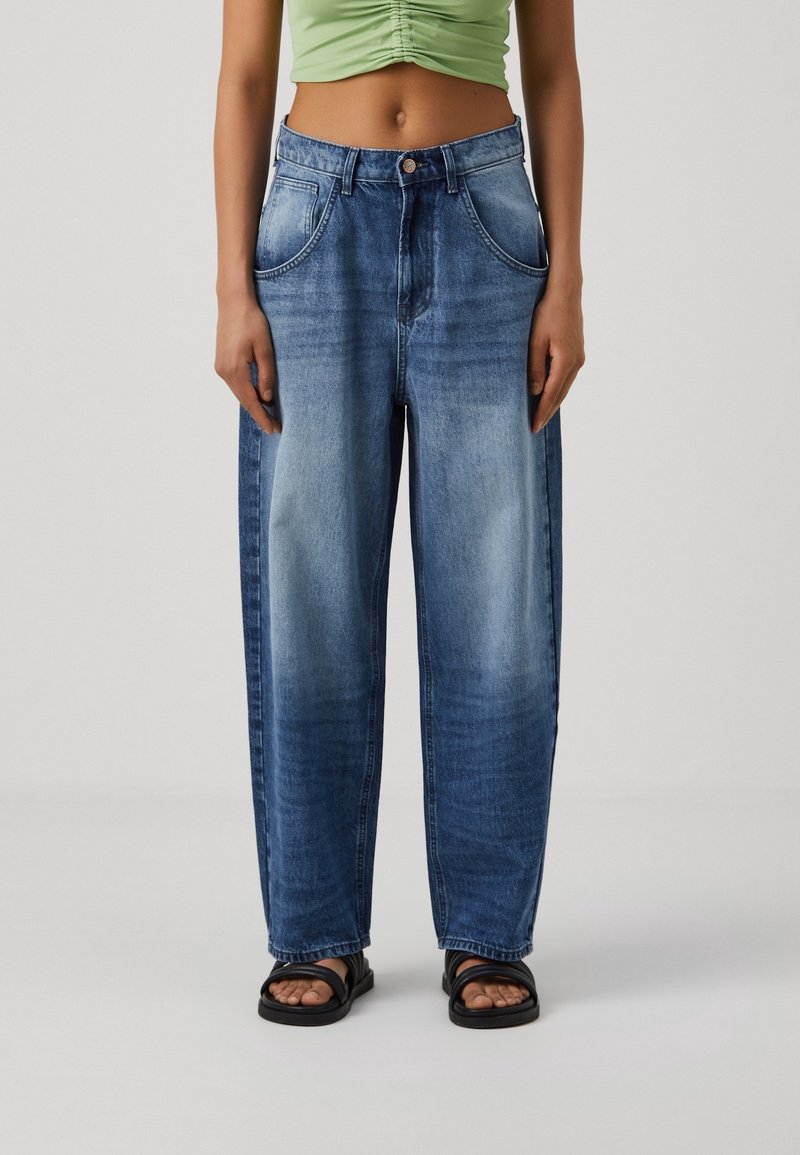 Personne portant un jean bleu délavé taille haute à jambes larges, un crop top vert froncé et des sandales noires sur un fond clair uni.