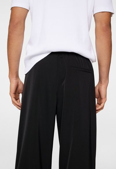 Black trousers with a smooth texture, featuring a single back pocket and a waistband with subtle pleats. Worn with a white shirt.