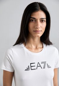Balta trumpa rankovėmis marškinėliai su juodu spausdintu logotipu, ant kurio užrašyta "EA7 EMPORIO ARMANI", su švelniu tekstūra ir apvaliu kaklu.