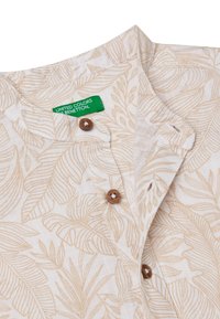 Camicia a maniche corte in tessuto leggero con motivo di foglie beige. Presenta bottoni in legno e un'etichetta verde con "United Colors of Benetton."