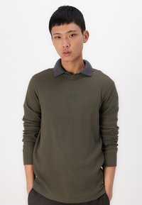 Tricou cu mânecă lungă, de culoare verde măslinie, cu guler polo de culoare gri închis, având un logo GAP brodat subtil pe piept. Textură netedă.