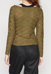 Maglietta a maniche lunghe verde oliva in tessuto texturizzato a coste. Presenta un ampio scollo e una silhouette aderente, abbinata a pantaloni neri.
