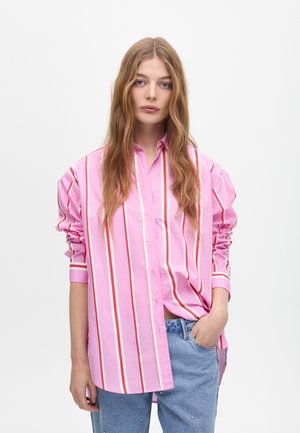 Camicia rosa con bottoni, a righe verticali in diverse tonalità, con maniche arrotolate e una vestibilità rilassata, abbinata a jeans azzurri chiari.