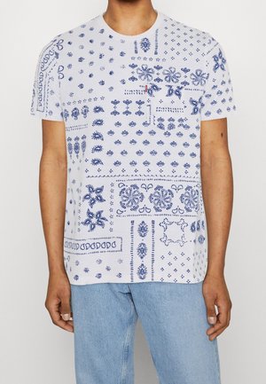 Homme portant un t-shirt blanc à manches courtes avec des motifs complexes de paisley et floraux bleus, associé à un jean denim bleu clair.