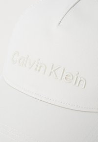 Casquette de baseball blanche en tissu texturé, avec un logo « Calvin Klein » en relief de couleur assortie et des détails de couture visibles.
