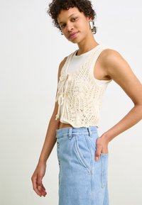 Häkelweste in Creme mit Bindeschluss, über einem figurbetonten weißen Top getragen, kombiniert mit einem hochtaillierten hellblauen Jeansrock mit Knöpfen.